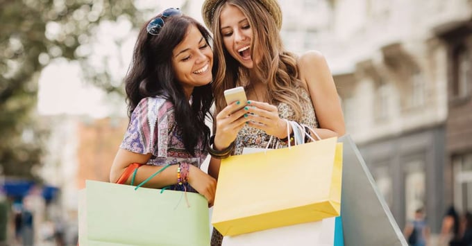 Two women holding shopping bags look at a phone to review their purchase.