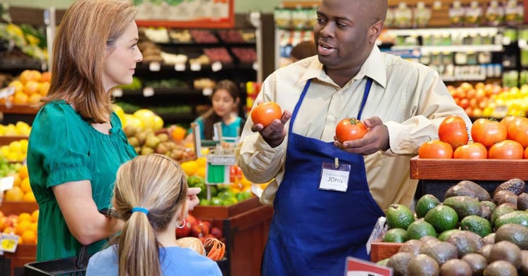 A grocery store worker holds up two tomatoes to show a female customer and her daughter substitute options for a grocery order.