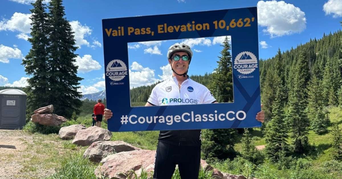 Eric Brounstein stands in cycling gear on top of a mountain holding a sign that says #CourageClassicCo