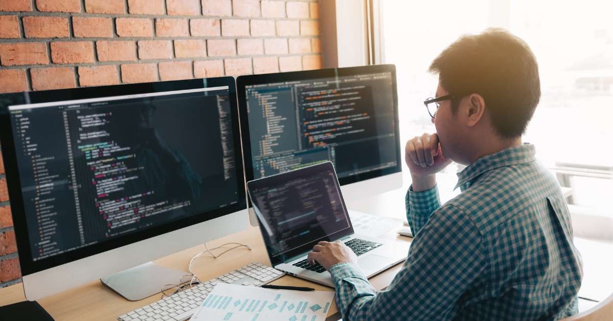 A man sits at a computer desk in front of two large monitors and types code into a laptop.