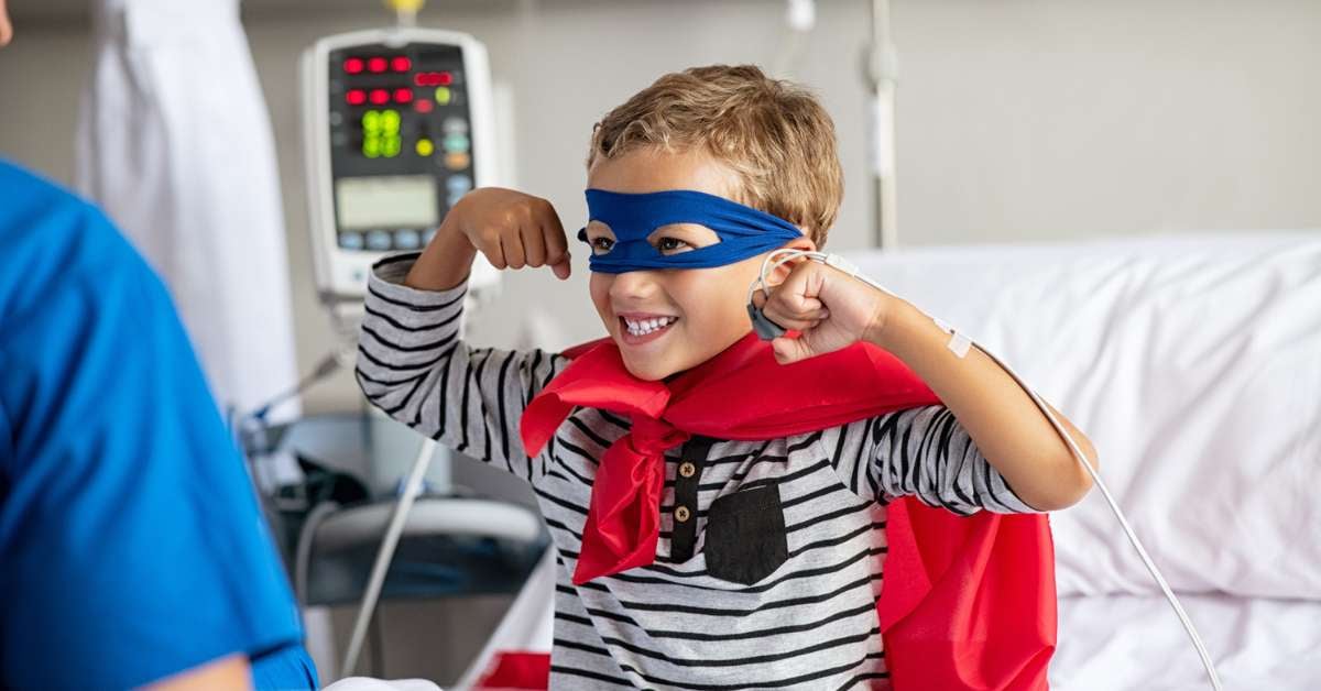 Little boy wears a super hero costume and flexes his muscles while he sits on a hospital bed.