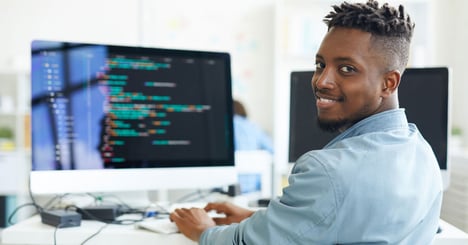 Man turns and smiles at the camera while running TestCafe application testing on a desktop computer.