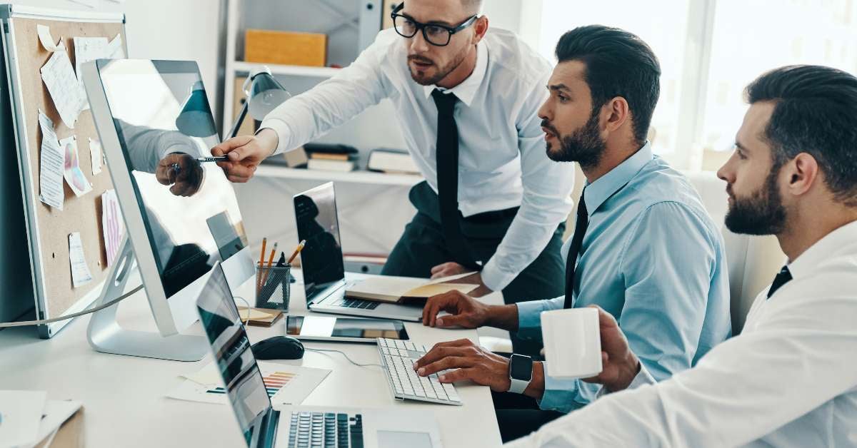 A group of coworkers sit at a desk and discuss new development tool options listed on a computer monitor.