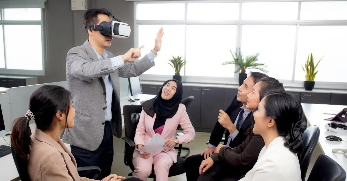 Man standing in a conference room wearing VR goggles surrounded by his female peers